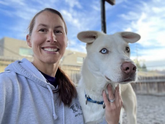 Smiling woman in a light gray hoodie poses with a pale dog that has blue eyes outdoors.