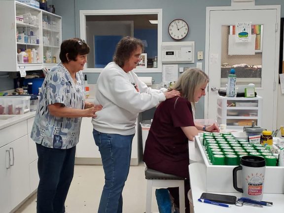 Three volunteers in a shelter kitchen packing meals for cats and dogs, with shelves of supplies in the background.
